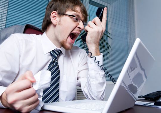 איש לבוש בחולצה לבנה עם עניבה מול מחשב נייד ועם הטלפון ביד צועק וכועס