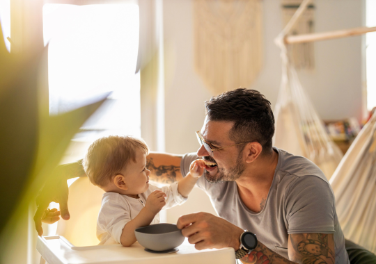 רקע של סלון של בית עם ערסל ועציץ, אבא מחייך אל הבן שלו התינוק שאוכל מקערת אוכל