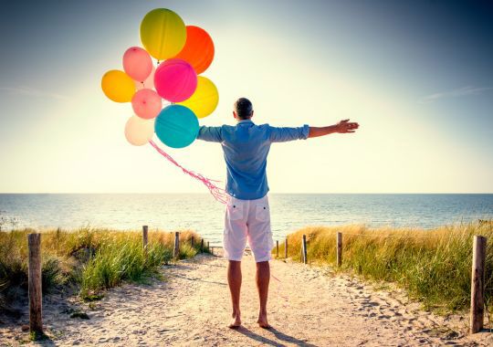 איש עם חולצה כחולה ומכנס לבן על שפת הים עם בלונים צבעוניים עם הליום ביד