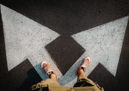 רואים מלמעלה רגליים של בחור עם נעלי אולסטאר כתומות כהות עומד עם הרגליים שלו על חיצים בכביש שכל רגל על חץ שפונה לכיוון אחר